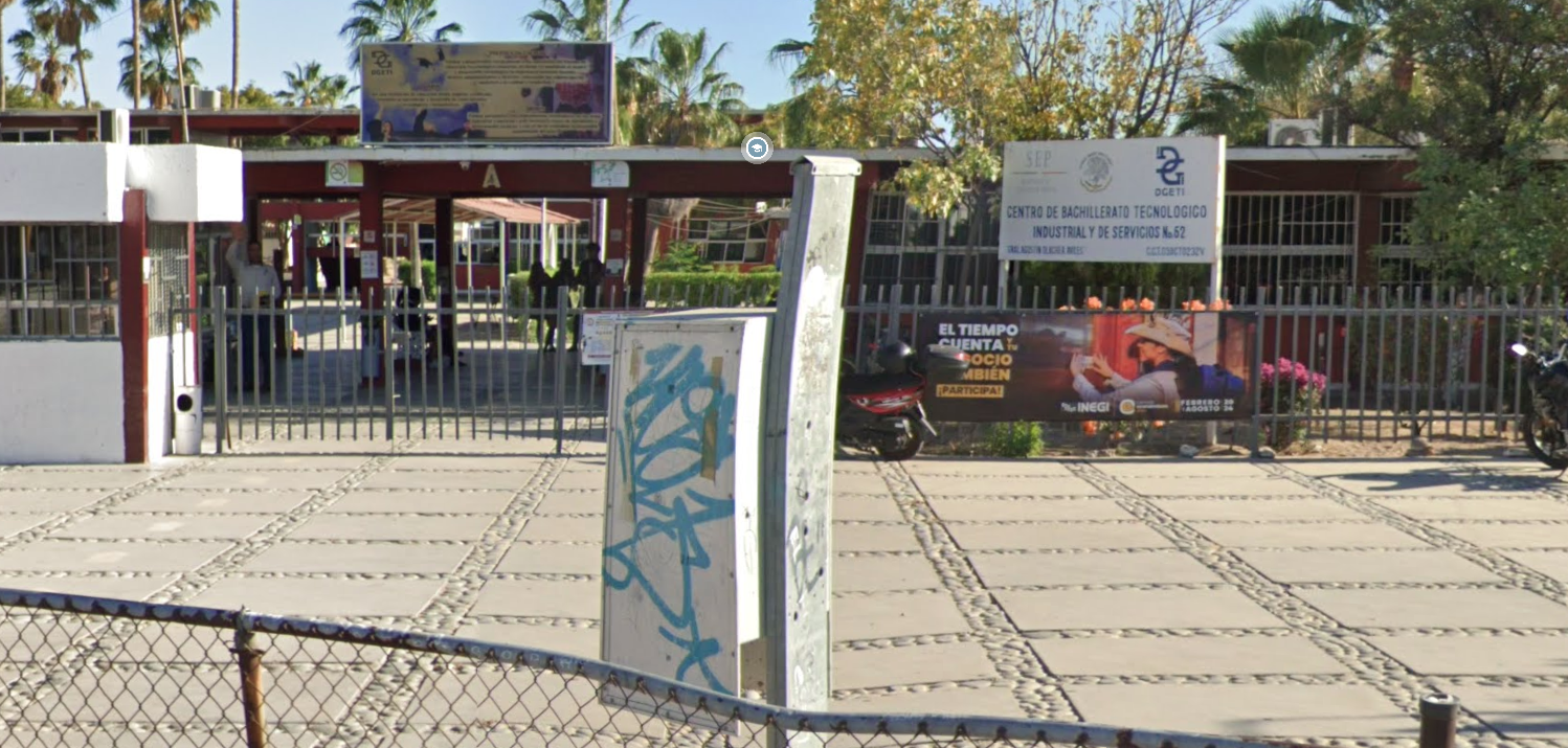 Fotografía en la que se muestra la explanada y al final se encuentra la entrada del plantel, conformada por una reja color gris. Arriba de la reja hay un cartel que en letras azules dice CENTRO DE BACHILLERATO TECNOLÓGICO INDUSTRIAL Y DE SERVICIO NÚMERO 62. 